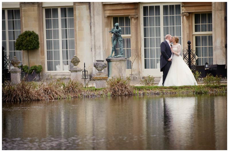 bride kisses her groom nest to water with woburn sculpture gallery in background
