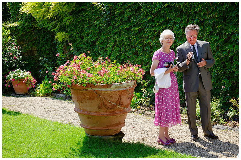 Man in suit holding video camera next to lady in Pink dress with white spots in gardens standing next to large terracotta palnt pot full of pink flowers