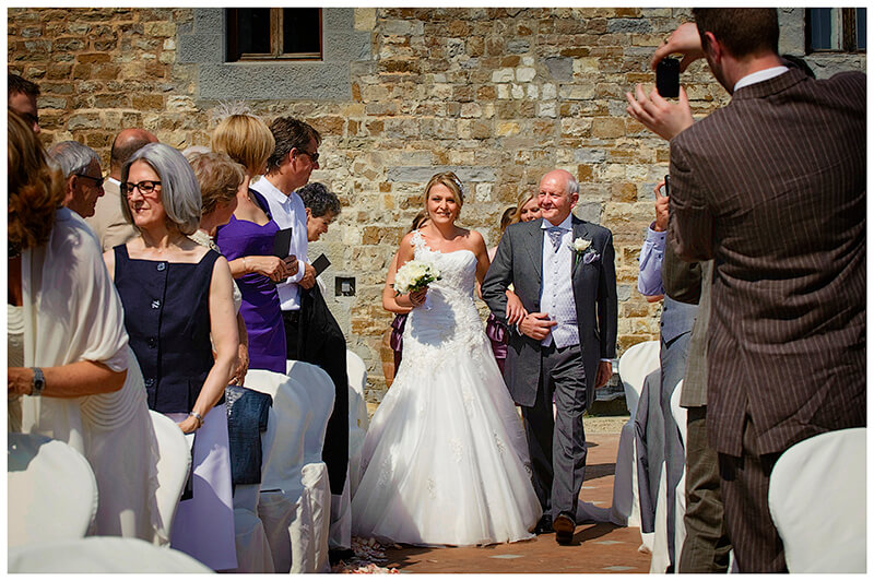bride and father walk down aisle as guests watch and take photos on phone