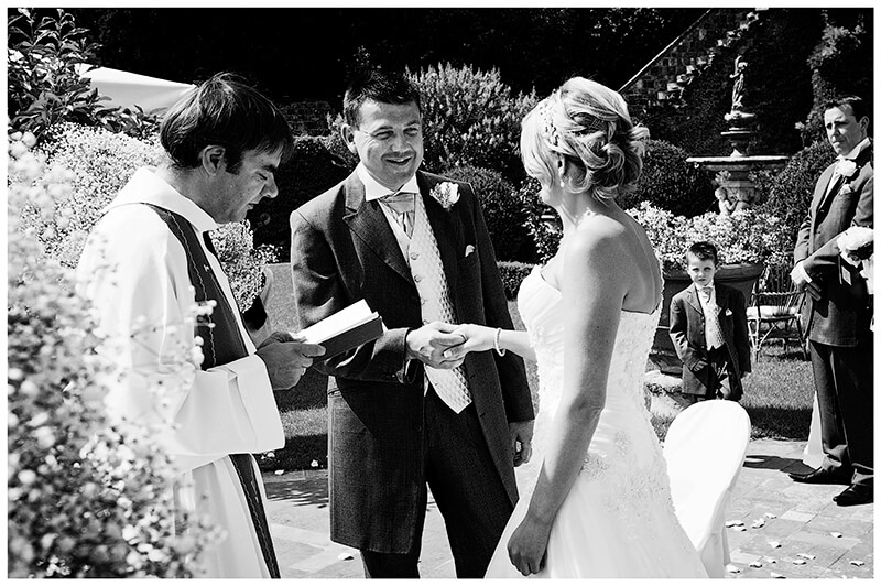 smiling groom holds his brides hand watched by bestman and paige boy in background