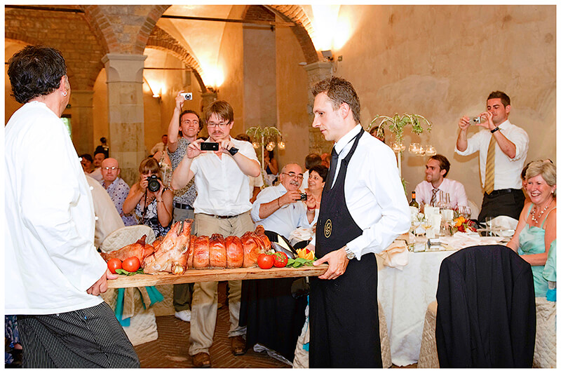 waiters carrying roasted pig on wooden tray being photographed by wedding guests