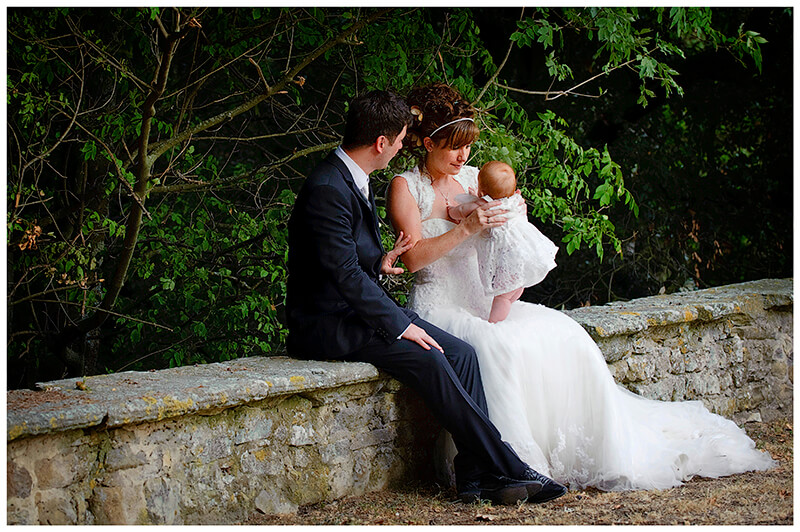 groom tenderly touches brides arm as she holds young flower girl in the gardens of Castel di Poggio 