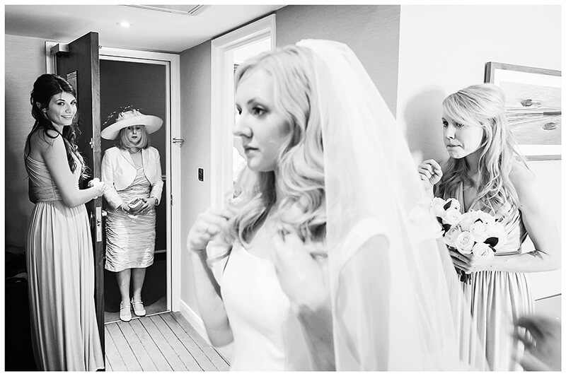 Bride adjusts veil mother standing in doorway bridesmaids watching 