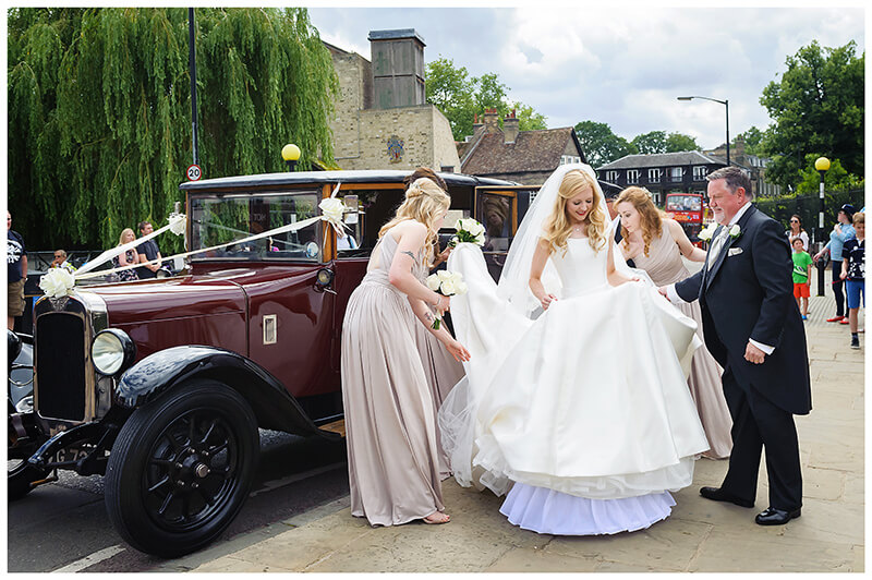 Bride help to get out of car by bridesmaids and father outside the entrance to Queens College wedding Cambridge