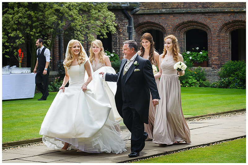 Smiling Bride looks at Father as Bridal Party walks through Queens College Cambridge Quad waiter in background