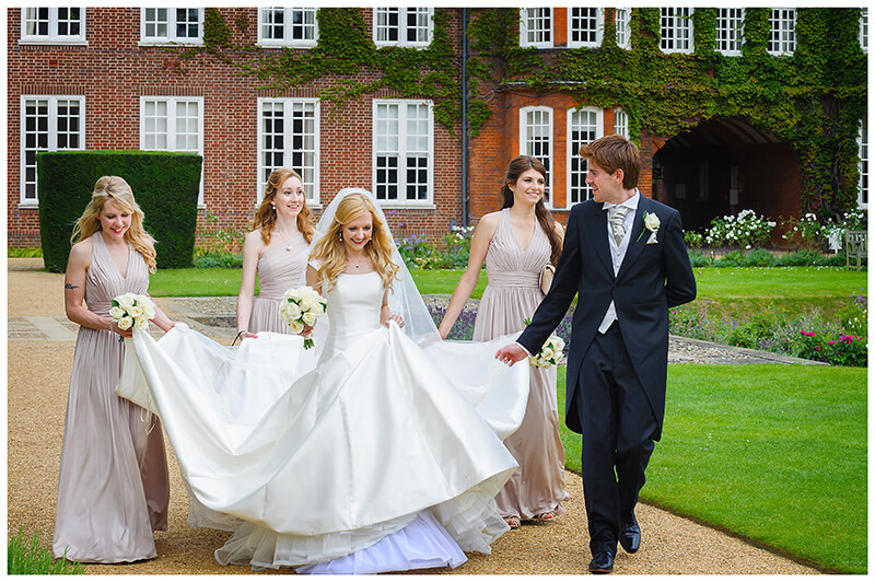Groom bridesmaids hold dress as bride walks through grounds of Newnham College Cambridge