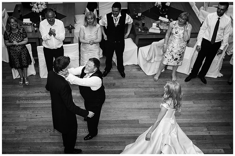 Father of brie clasps grooms face on dance floor as bride and guests watch