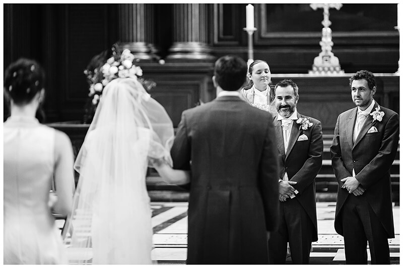 smiling groom watches his bride walk down aisle of Trinity College Chapel