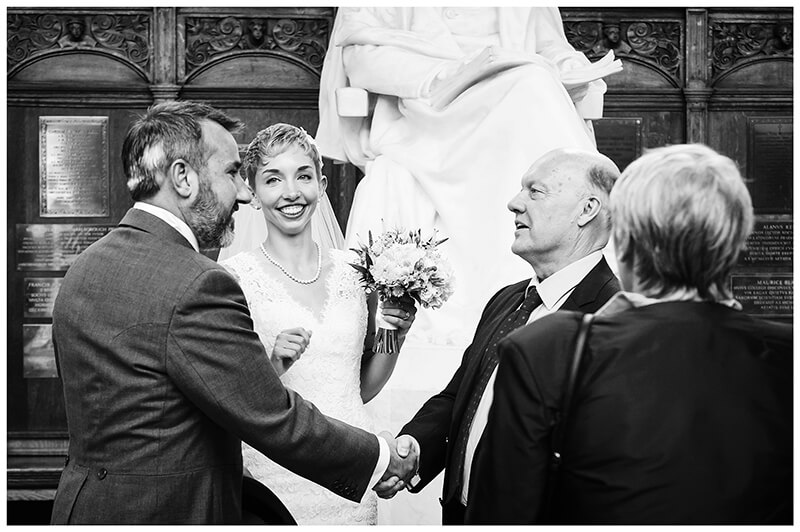bride smiles as groom shakes hands with guests at Trinity College wedding