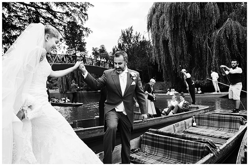 bride being helped by groom and bridesmaid to get on punt watched by tourists on bridge