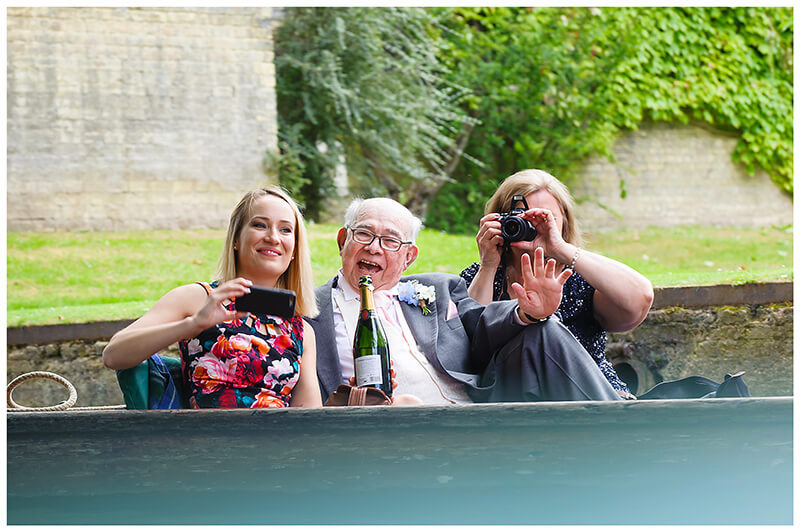 three wedding guests on punt taking photo holding champagne bottle and waving