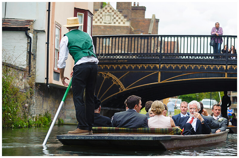 lady standing on bridge watches as wedding party passes underneath on punts on river in Cambridge