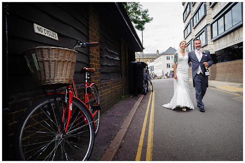 bride and groom walking along street in Cambridge passing red bike with NO Cycles sign