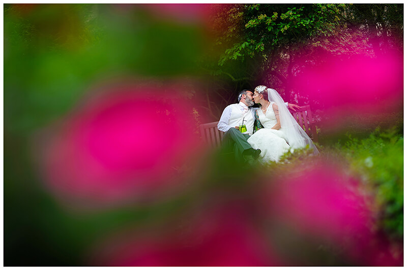 bride and groom kiss sitting on bench surrounded by purple flowers