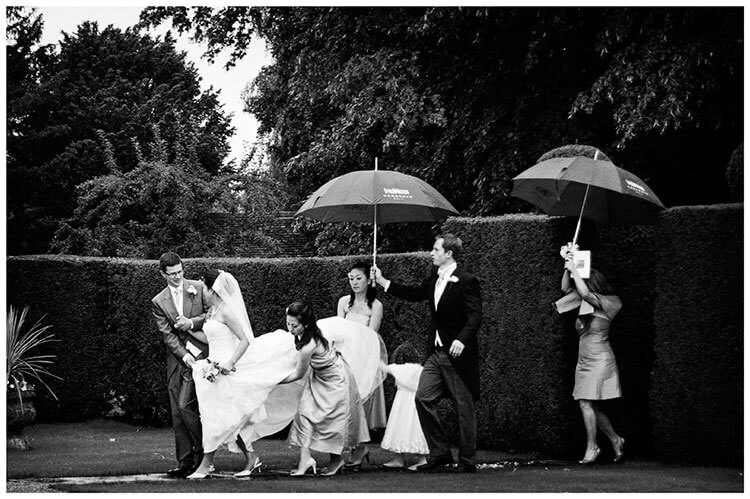 hengrave hall wedding party walking under umbrellas bridesmaids lifting brides dress