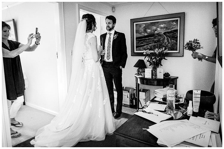 Bride talks to brother in hallway as make up artist takes photo of the back of the brides veil in hair whilst brides mum enters holding bouquet. favourite wedding photography of 2018