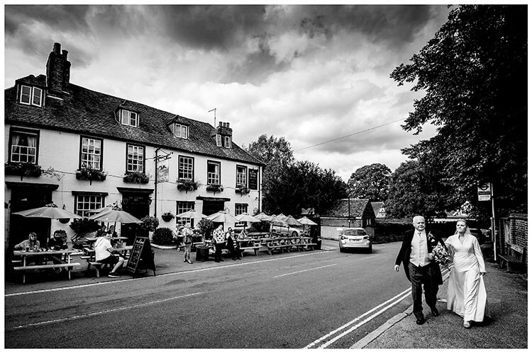 Bride and her father walking to the church with people in the pub garden looking on