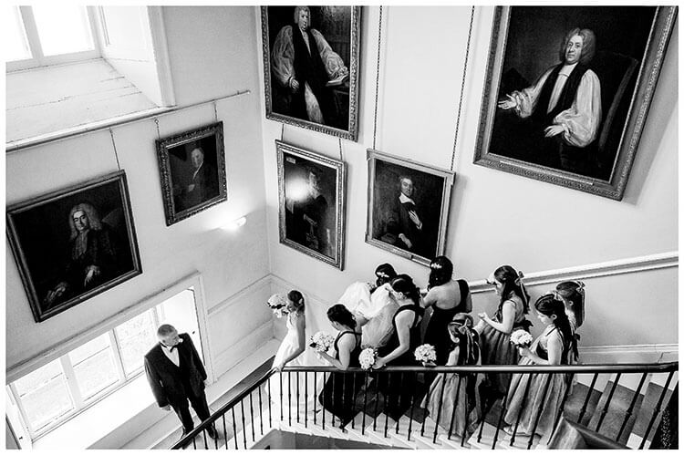 Father of Bride waits on stairs for the Bridal party to catch up, grand portraits on walls