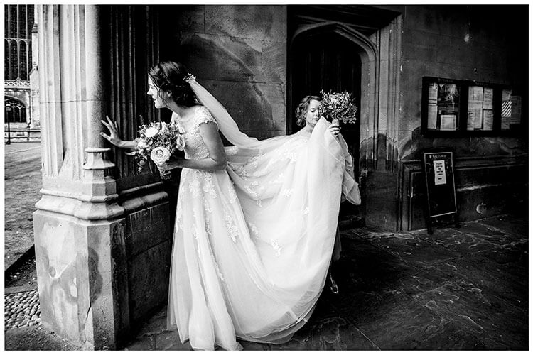 Bride checking all guests have arrived for wedding ceremony at Kings College Cambridge as Bridesmaid lifts back of brides dress