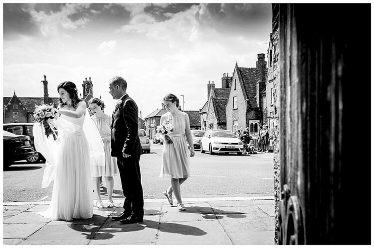 Bride adjusts viel at enterance to Ramsey Cambrideshire church watched by Father and Bridesmaids