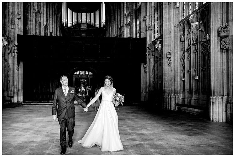 Smiling Bride groom walk hand in hand out of Kings College chapel after their wedding ceremony followed by all their guests