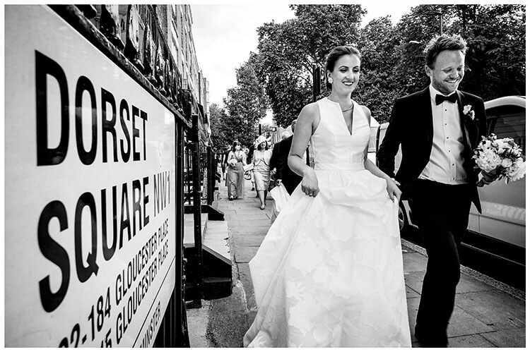 Smile bride and groom walking along street pass sign for Dorset Square in London followed by guests, bride lifts front of dress groom carrying bride bouquet during London favourite wedding photography 2018