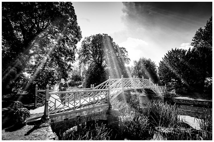 Bride and groom standing on bridge, in the grounds of Island Hall wedding venue in Cambridgeshire, drink Champagne as wonderful rays of light shine down on them favourite wedding photography 2018